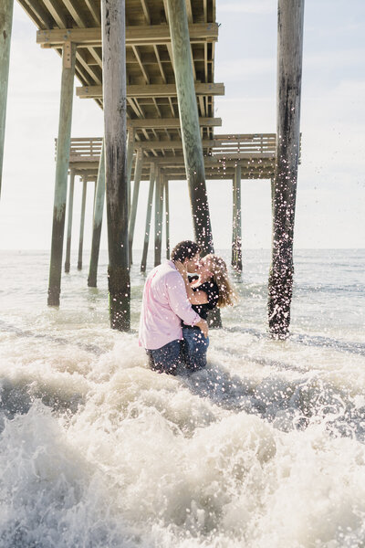 Engaged couple kisses under the fishing pier in Ocean City, NJ as the waves crash around them