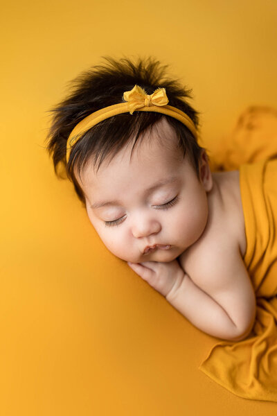 Beautiful baby girl with a full head of hair posed and sleeping on a yellow backdrop.