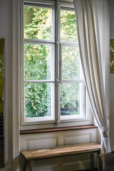 Curtains hand in front of large, white window with a small wooden bench beneath it.