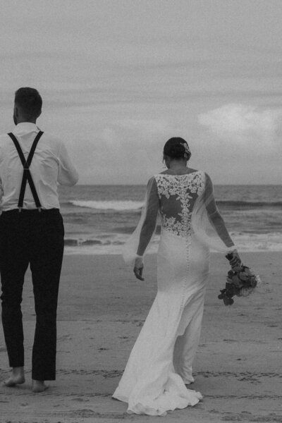 Newlyweds walking on the beach, groom in suspenders, bride in a lace-back gown.