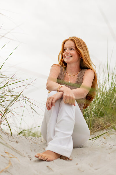 Senior girl with red hair sitting on sandy beach surrounded by tall grass - link to senior photography