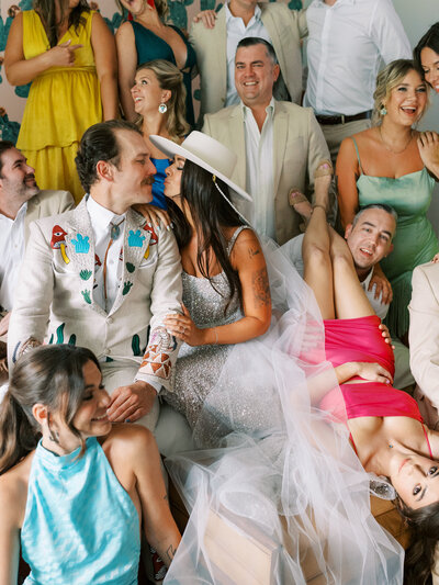 groom in printed suit kissing bride in hat on a hotel room bed with all of their wedding party around them