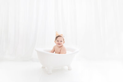A toddler girl sits in a small white tub in a yellow bow in a studio