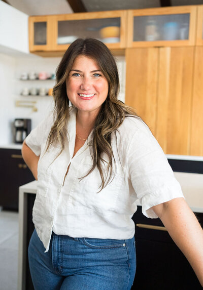 Lead interior designer, Michelle, wearing a white linen top and jeans standing in a kitchen with wood and black cabinets