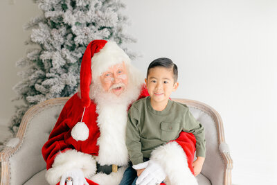 Child in dark green shirt sits on an Indianapolis Santa's lap and smiles for photos with santa
