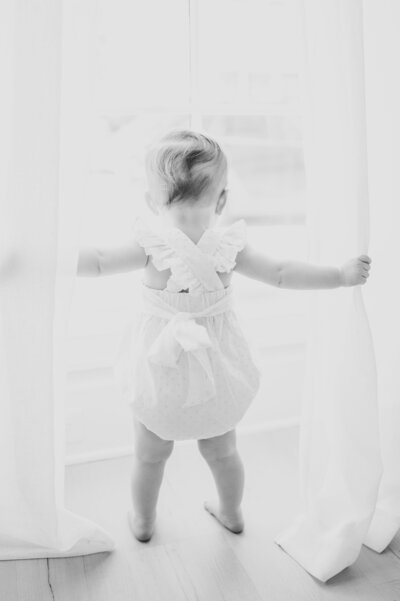 Little girl in ruffled romper looks out of window holding curtains during photo session in Raleigh NC