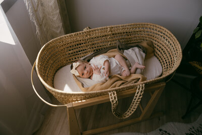 Sweet newborn photography shoot of a baby girl in a bow and white vintage looking onesie looking up at the camera,  laying in a basket with blankets under her captured by Morgan Ashley Lynn Photography in Lake Country, WI