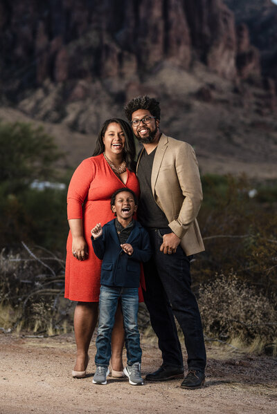 Photo of a happy black family with mom a dad and a young son.  All are dressed nicely in front of a mountain with big smiles