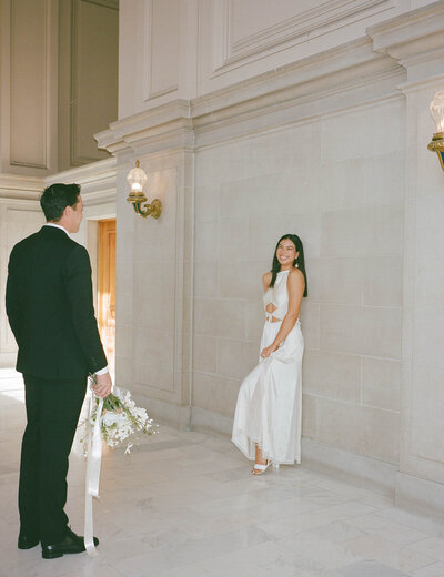 Person in a black suit admiring their parnter in a white wedding dress leaning against the wall at City Hall