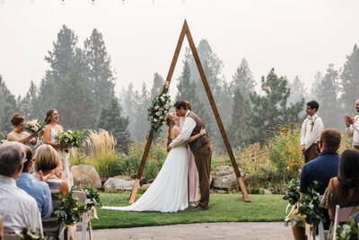 A triangle arbor decorated with greenery and white and pink roses frames a redheaded bride wearing a strapless gown while her groom, in brown vest and slacks kisses his bride right after their Aspen Lakes Golf Course Wedding Ceremony in Sisters, Oregon. | Erica Swantek Photography