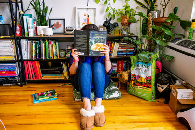 Woman reading book, The Land Where Lemons Grow, in her apartment surrounded by house plants