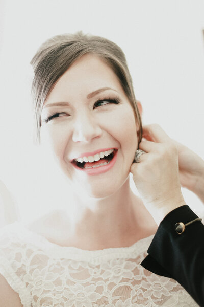 Bride smiles while putting on earrings before wedding