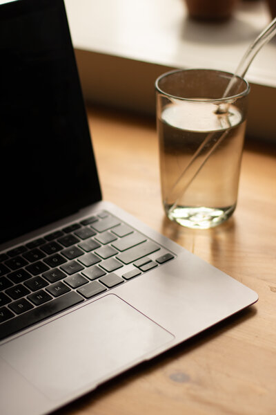 Portion of macbook and glass of water on a wooden table lit by natural light