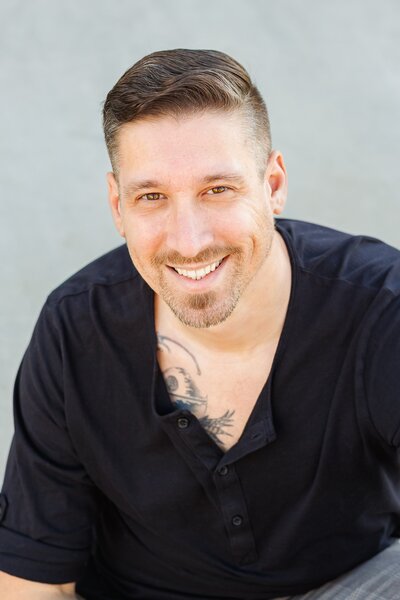 Professional headshot of man with brown hair and a beard and tattoo peeking through black shirt