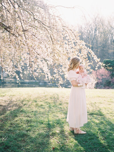 Mom with blonde hair in white dress holds baby girl in front of cherry blossom tree.