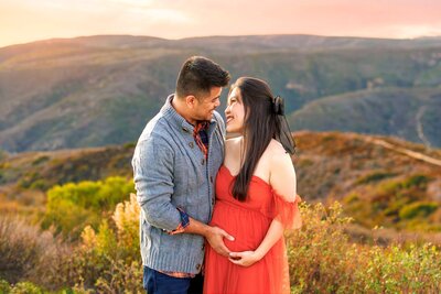Couple looking at each other and holding pregnant belly in front of mountain views at Top of the World trails in Laguna Beach