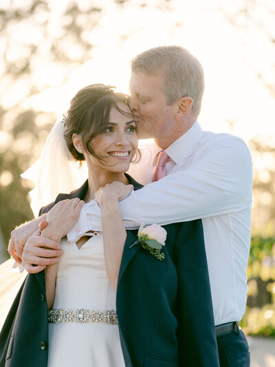 bride wearing groom's jacket as he hugs her from behind and kisses her on the forehead during golden hour romantics at el encanto a belmond hotel in santa barbara, ca captured by magnolia west photography los angeles wedding photographers