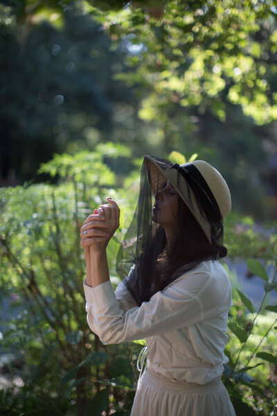 woman holding a bee in a hat with a sheer bee veil, green foliage in the background