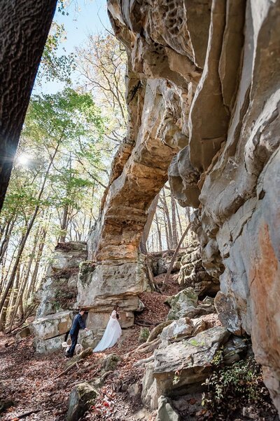 A bride and groom and their tan and white dog walk  in glowy sunlight under a huge light colored sandstone arch.  Sunlight streams through the green leave of trees during their destination elopement with their photographer in Tennessee.
