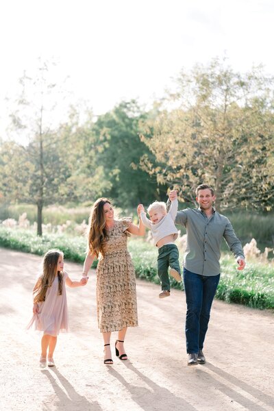 Family of mom and dad with young daughter walking outside with son swinging in the air holding parents hands