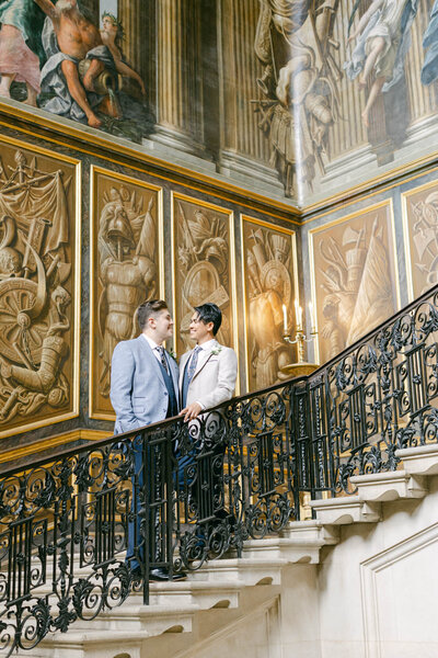 at a luxury gay wedding two grooms hold hands on the staircase at Hampton court palace with the mural staircase walls as their backdrop