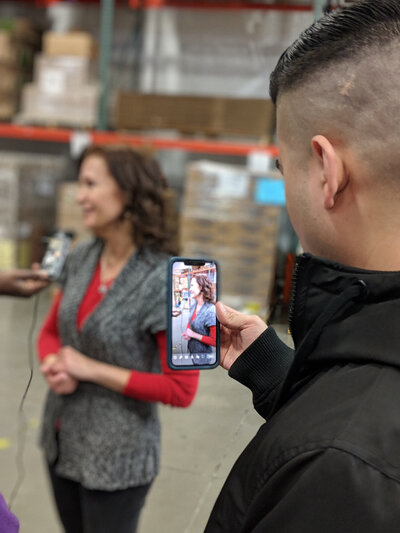 A man holds up his phone to photograph a woman who’s standing in the background.