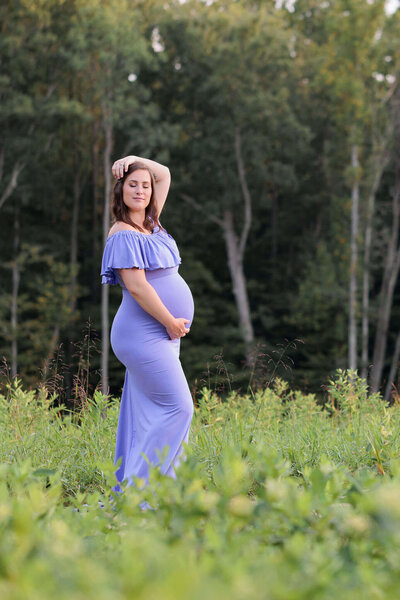 A mom-to-be wearing a purple dress and holding her belly with her eyes closed at a park in Northern Virginia.