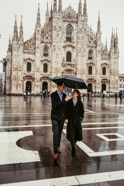 Couple walking in front of Duomo church in Milan Italy while holding an umbrella under the pouring rain