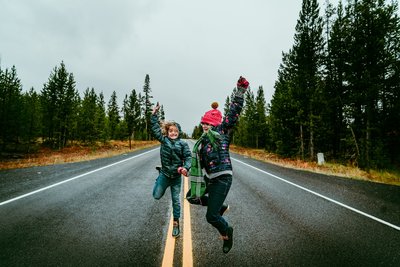 Two kids exuberantly jump in the air on an empty road, lined by pine trees
