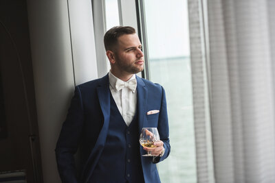 Groom in a blue suit looking out window on wedding day sipping whiskey.