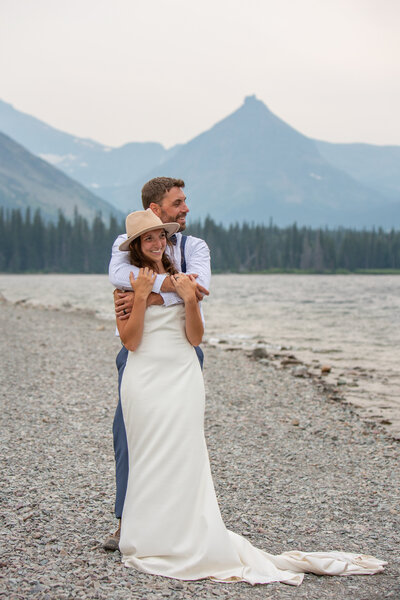 A bride in a white dress stands with her groom with his arms wrapped around her shoulders on a lakeshore in Montana