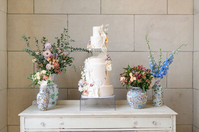 White 5 Tiered wedding cake with a clear spacer used as the middle tier decorated with florals sitting on a table in between two small bouquets inside painted vases.