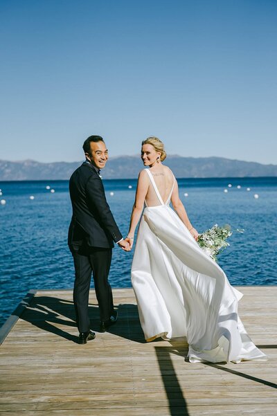 bride and groom looking over their shoulders right after thier first look on a dock on west shore of Lake Tahoe. Wind blowing in bride's dress, holding her bouquet.
