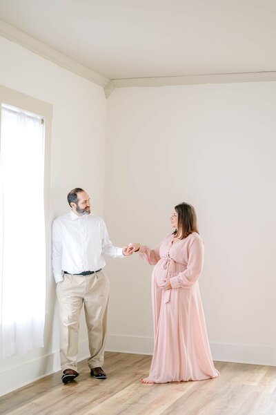 A husband takes his wife hand and gently guides her towards him as she cradles her baby bump in a pink flowy dress