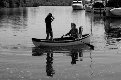 Behind the scenes black and white image Mark Maryanovich standing in row boat while photographing musician Stephen Fearing  who holds oars