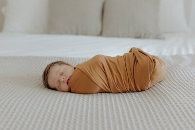 A newborn sleeping on a bed in a brown swaddle.