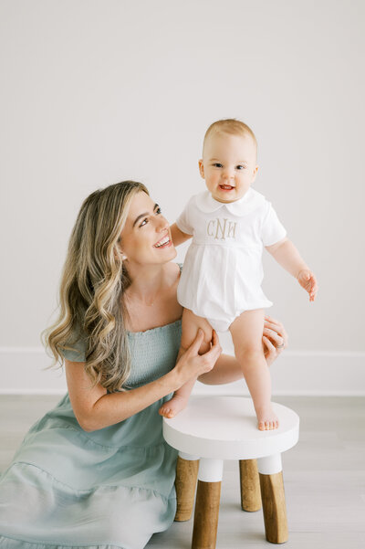 One year old baby boy in white monogrammed romper stands on stool with mom holding his legs during photo session by Worth Capturing Photography