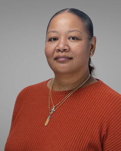 A person stands on stone steps in front of a large stone building, holding a bright red coat. They are wearing a white dress and red shoes. Trees are visible in the background.