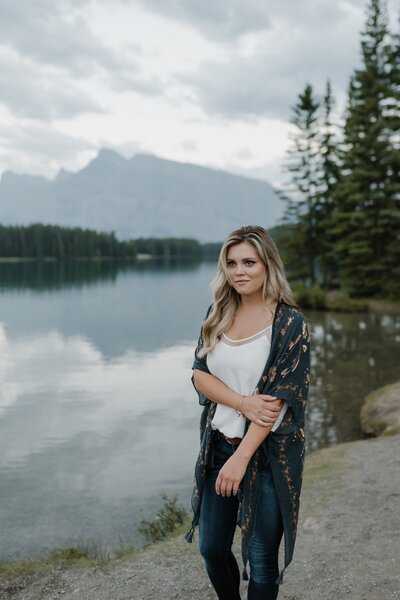 Couples and Lifestyle photographer smiles softly at the camera  with one arm holding the other while standing in front of a mountain lake