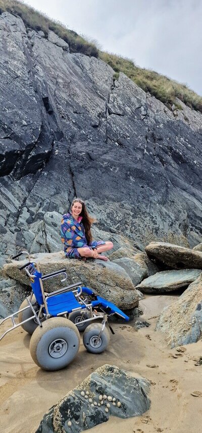 Bethany Handley smiling while sitting barefoot on large coastal rocks, with a beach wheelchair resting nearby on the sand.