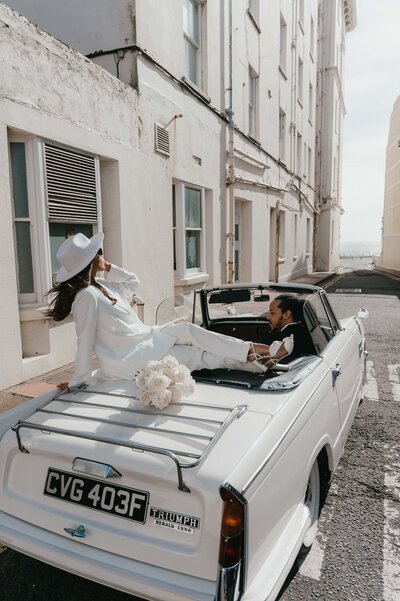 Shany sitting on top of white cabriolet with bridal bouquet and tony driving the car in the streets of London after their elopement.