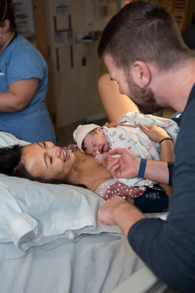 A mother is laughing with her brand new baby on her chest as her partner smiles and holds the baby's tiny hands at UW Medical Center at Montlake, WA.
