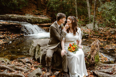 Bride and Groom sit on a rock together while he holds her face. They sit in front of a waterall during their Pennsylvania fall elopement at Hickory Run State Park.