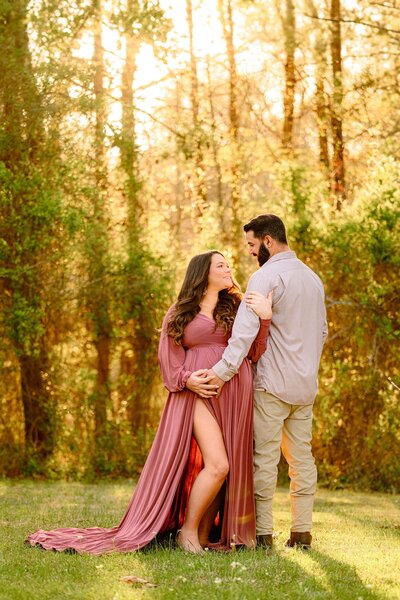 Pregnant mother stands with a glow of the sun behind her and a rose pink dress with her hand and her husband's hand on her belly each looking towards each other during their maternity session
