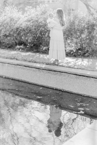 A black and white image of a mother holding her baby girl with her reflection in a  reflection pool