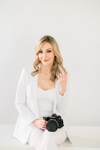 woman sitting on a white desk wearing a white suit, smiling and holding her camera