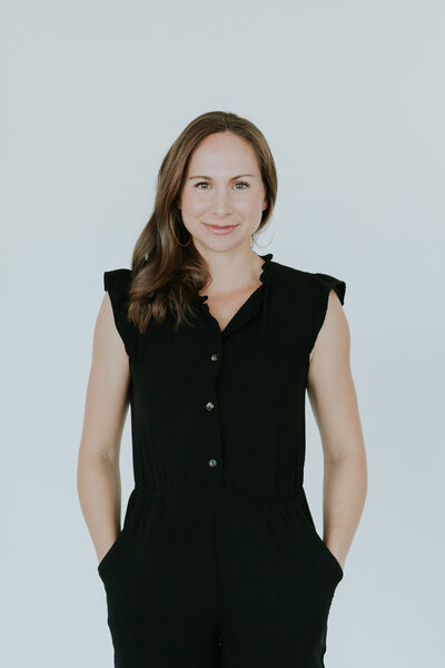 Professional headshot of Sarah Ingle of Nest Analytics with a closed mouth smile, with hands in pockets, wearing black jumpsuit in front of white background at The Frame Charlotte