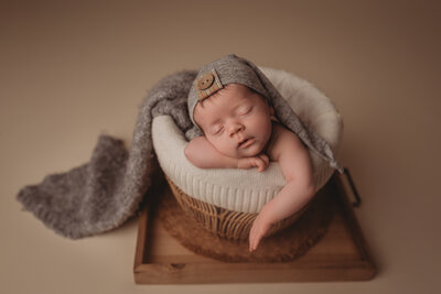 Newborn baby boy asleep at Woodstock, GA newborn portrait studio posing with chin on hands in basket wearing gray sleepy cap.