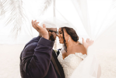 A bride and groom kissing under a veil on the beach.
