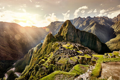 A breathtaking view of Machu Picchu at sunrise. The ancient Incan city is nestled among lush, misty mountains, with terraces and stone structures clearly visible. Sunlight illuminates the scene, casting a warm, golden glow over the landscape.